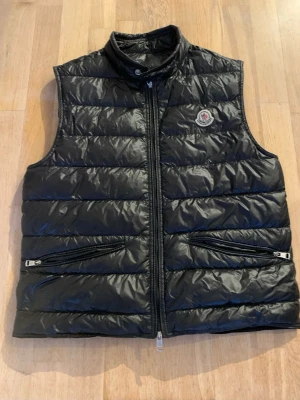 Svart dunväst från Moncler - Snygg svart dunväst från Moncler med glansig finish och klassisk logga på bröstet. Västen har hög krage, två dragkedjeförsedda fickor och quiltad design. Perfekt för lager-på-lager och streetstyle.