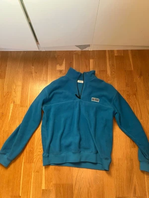 Blå fleece från Junkyard Urban Tears - Blå fleece-tröja från Backyard med Urban Tears-märke på bröstet. Tröjan har halv dragkedja, hög krage och ribbade muddar. Perfekt för kyliga dagar och enkel att slänga på sig över en t-shirt. Materialet är mjukt och värmande fleece.