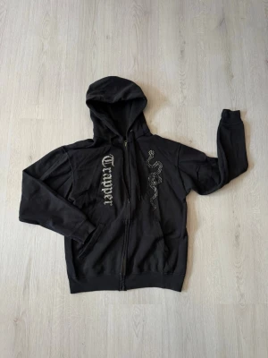 Rhinestone Hoodie - Svart hoodie med huva och dragkedja framtill. På ena sidan står det 'Trapper' i gotisk stil och på andra sidan finns ett ormmönster i silvriga nitar. Klassisk passform och känguruficka framtill. Perfekt för en streetwear-look.