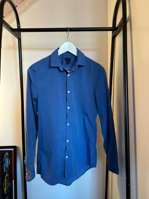 Blå skjorta med knappar - Snygg blå skjorta med klassisk krage och vita knappar. Skjortan har lång ärm och rak passform, perfekt för en stilren look. Tillverkad i ett lätt och bekvämt material som passar året runt.
