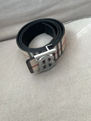 Rutmönstrat Burberry bälte i skinn - Säljer ett klassiskt Burberry bälte i skinn med det ikoniska rutmönstret i beige, svart och rött. Bältet har ett snyggt silverfärgat B-spänne som ger en lyxig touch. Perfekt accessoar för att lyfta vilken outfit som helst.