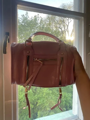 Rosa handväska med dragkedjor - Snygg ljusrosa handväska i glansigt syntetmaterial med två dekorativa dragkedjor framtill och ett handtag upptill. Väskan har guldfärgade detaljer och en kompakt, boxig form som ger en cool vibe. Perfekt för att lyfta din outfit.