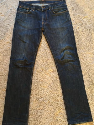 Mörkblå jeans från Nudie Jeans - Säljer ett par mörkblå jeans från Nudie Jeans med klassisk femficksmodell och raka ben. Jeansen har snygga slitningar vid knäna och bakfickor med Nudie Jeans. Tillverkade i denim av hög kvalitet. Skriv gärna om frågor uppstår! 😁
