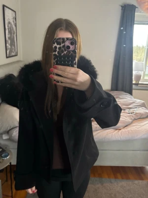 Svart kappa med pälsluva - Säljer en svart kappa med stor luva och pälsdetalj runt huvan. Kappan har dubbelknäppning framtill och vida ärmar för en trendig look❤️