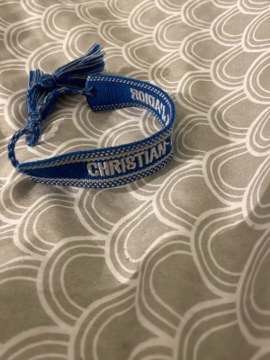 Blått Christian Dior tygarmband - Snyggt blått armband från Christian Dior i vävt tyg med vita detaljer och tofsar. Armbandet har märkets logga broderad i vitt och justerbar knytning för perfekt passform. Perfekt accessoar för att lyfta din outfit.