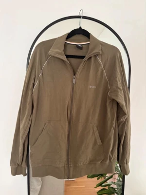 Beige zip-hoodie från BOSS - Snygg beige hoodie från BOSS med dragkedja framtill och två fickor. Tröjan har en stilren design med diskret BOSS-logga på bröstet och vita detaljer längs axlarna. Tillverkad i mjukt bomullsmaterial och har en klassisk krage utan huva.
