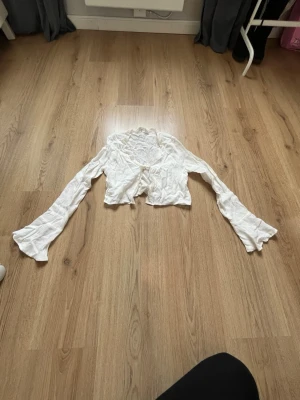 Vit blus med volangkant och knyt - Söt vit blus med långa ärmar, volangkant längs urringningen och knyt framtill. Blusen är kortare i modellen och har en lätt ribbad struktur. Perfekt för en trendig och romantisk look.