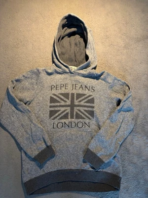 Grå hoodie från Pepe Jeans London - Snygg grå hoodie från Pepe Jeans London med stort Union Jack-tryck och text på bröstet. Kashmir, bomull,ull. Storlek 164 14år. Använts få gånger.