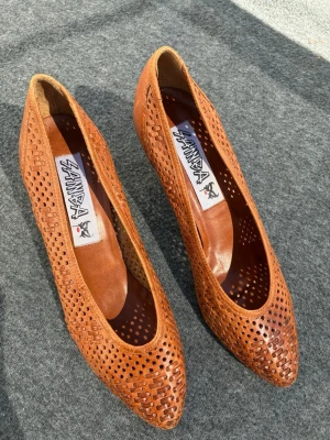 Bruna pumps i läder - vintage - Bruna pumps i äkta läder med perforerat mönster och spetsig tå. Skorna har medelhög klack och flätade detaljer framtill som ger en unik look. Perfekt för dig som gillar vintagevibbar! Klacken är nyklackad. Mycket bra skick!