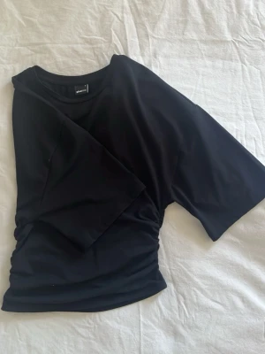 Svart t-shirt med rynkade detaljer - Svart t-shirt från Ginatricot med korta ärmar och rynkade detaljer i sidan. T-shirten har en loose passform och är tillverkad i mjuk bomull, perfekt för en avslappnad och trendig look.