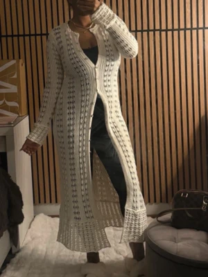 Lång vit stickad cardigan - Lång, öppenstickad vit cardigan med knappar framtill och långa ärmar. Cardiganen har ett luftigt mönster och är figurnära upptill med utsvängd nederdel. Perfekt för lager-på-lager och ger en chill vibe.