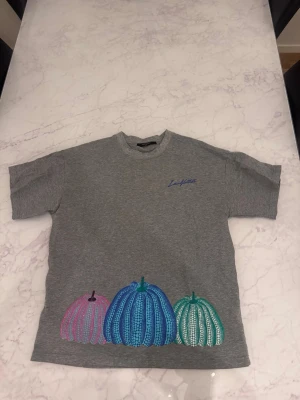 Grå t-shirt med färgglada pumpor - Grå t-shirt med korta ärmar och tre broderade pumpor i grönt, orange och rosa längst ner på ryggen. T-shirten har en klassisk passform och är tillverkad i mjuk bomull. Perfekt för dig som gillar unika detaljer och färgglada motiv.