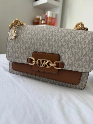 Michael Kors axelväska med kedja - Snygg axelväska från Michael Kors i beige och brunt konstläder med MK-logga och guldfärgad kedja. Väskan har ett mönstrat yttre, dekorativt spänne framtill och en söt blomcharm. Perfekt för dig som vill ha en trendig och stilren accessoar.