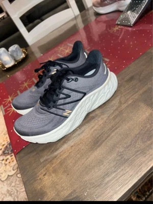 Grå New Balance Fresh Foam sneakers - Snygga grå sneakers från New Balance med Fresh Foam-mellansula för extra komfort. Ovandelen är i meshmaterial med svarta detaljer och snörning. Skorna har en chunky vit sula och diskret guldigt N-logga på sidan. Perfekta för en sportig och trendig look.
