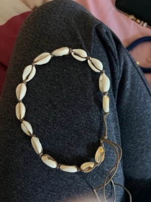 Snäckhalsband med justerbart band - Ett snyggt halsband med vita snäckor trädda på ett brunt justerbart band. Perfekt accessoar för en bohemisk eller strandinspirerad stil. Snäckorna är ovala och har en naturlig glans. Inget märke syns på halsbandet.