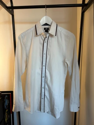Vit skjorta med svarta detaljer H&M - Vit skjorta från H&M.  Klassisk krage och långärmad modell i bomull. Perfekt för dig som gillar stilrena plagg med en twist.