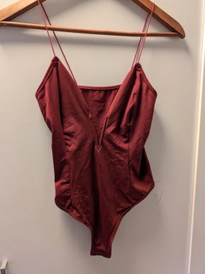 Vinröd body med smala axelband - Säljer en vinröd body med djup V-ringning och smala axelband. Bodyn är tillverkad i ett mjukt, stretchigt tyg som sitter snyggt mot kroppen. Perfekt att styla med jeans eller kjol för en trendig look.Liten i storlek 