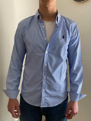 Blårandig skjorta från Ralph Lauren - Snygg skjorta från Ralph Lauren med blå och vita vertikala ränder. Klassisk krage, långa ärmar och knäppning framtill. Liten broderad logga på bröstet. Perfekt för en stilren look.