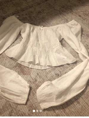 Vit offshoulder blus med puffärm - Romantisk vit blus i linne med offshoulder-modell och långa puffärmar. Blusen har en lätt rynkad midja och volang nedtill för en feminin siluett. Perfekt att styla med jeans eller kjol för en trendig look.