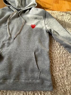 Grå hoodie  - Grå hoodie med huva och dragsnören. Klassisk känguruficka framtill och det ikoniska röda hjärtat med ögon broderat på bröstet. Mjuk bomullsblandning som är skön att ha på sig. Storlek M, passar dig mellan 173-180 cm lång 