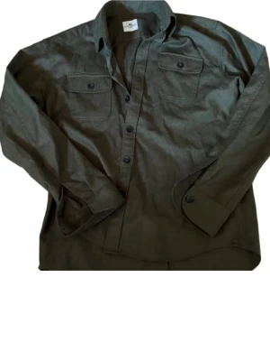 Olivgrön skjorta med bröstfickor - Stilren olivgrön skjorta med två bröstfickor och knappar framtill. Skjortan har lång ärm och klassisk krage, perfekt för en avslappnad men ändå snygg look. Tillverkad i ett mjukt bomullsmaterial som känns bekvämt hela dagen.