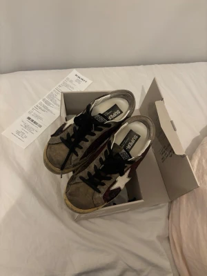 Golden Goose Superstar sneakers - Snygga Golden Goose Superstar sneakers i beige mocka med svarta skosnören, vit stjärndetalj på sidan och vinröda glitterpaneler. Klassisk låg modell med slitna detaljer för en cool och avslappnad look. Som ni ser på bilderna finns det defekter/slitningar vid de vinröda glittret. Perfekta skor annars. Original pris 5299kr och kvitto tillkommer samt låda och påse. 