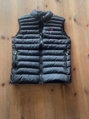 Svart dunväst från Polo Ralph Lauren - Snygg svart dunväst från Polo Ralph Lauren med horisontella sömmar, dragkedja framtill och två fickor med dragkedja. Klassisk röd logga på bröstet och hög krage. Perfekt för lager på lager och streetstyle.