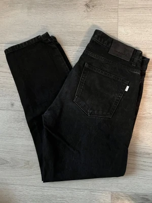 Svarta raka jeans från Woodbird - Säljer ett par klassiska svarta jeans från Woodbird med rak passform och fem fickor. Jeansen har en diskret logotyp på bakfickan och är tillverkade i slitstark denim. Perfekta för en clean och tidlös look.