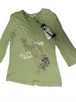 Ljusgrön topp  - Ljusgrön blus från By S.Oliver med v-ringning och dekorativa stenar vid halsen. Toppen har trekvartsärm och ett blommigt tryck i svart och vitt framtill. Tillverkad i mjuk bomull för en bekväm känsla.