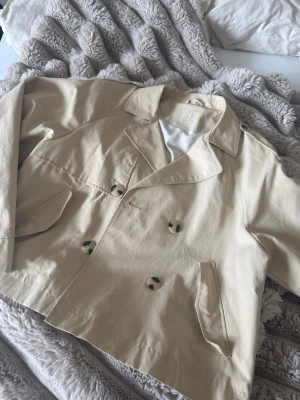 Kort beige trenchcoat från ZARA - Snygg kort trenchcoat från ZARA i beige med dubbelknäppning och klassisk krage. Jackan har två snedställda fickor framtill och stora knappar i sköldpaddsmönster. Tillverkad i ett slätt, lätt material som ger en clean look.