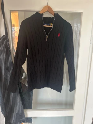 Svart kabelstickad tröja Polo Ralph Lauren - Kabelstickad svart tröja från Polo Ralph Lauren med halv dragkedja och röd broderad logga på bröstet. Tröjan har hög krage, långa ärmar och ribbade muddar. Perfekt avslappnad och klassisk stil.