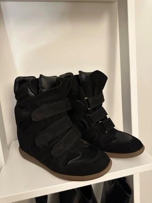 Svarta Isabel Marant sneakers med kardborre - Snygga svarta sneakers från Isabel Marant med högt skaft och flera breda kardborreband. Skorna har en mix av mocka och läderdetaljer samt en brun gummisula. Perfekta för dig som gillar streetstyle och vill ha något unikt till din outfit. Osäker om dom är äkta. Köpte på Vinted för 1000kr