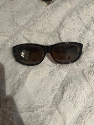 Svarta solglasögon från Polaroid - Snygga svarta solglasögon från Polaroid med klassisk cat eye-form och bruna glas. Ramen är i blank plast och har en diskret logga på sidan. Perfekta för att lyfta din stil under soliga dagar.