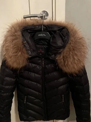 Svart pufferjacka med fuskpäls - Snygg svart pufferjacka från Chelsea med stor luva och bred, fluffig fuskpäls i brunt runt huvan. Jackan har dragkedja framtill, två fickor med dragkedja och quiltad design. Perfekt för kalla vinterdagar och riktigt trendig look.