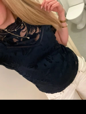 Mörkblå spetsblus med hög krage - Snygg mörkblå blus i spets med hög krage och transparenta partier. Blusen har korta ärmar och ett elegant blommönster i spetsen. Perfekt för dig som vill ha en trendig och feminin topp med fina detaljer.