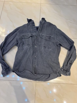 Mörkgrå jeansjacka med bröstfickor - Snygg mörkgrå jeansjacka med två stora bröstfickor och knappstängning framtill. Klassisk rak passform och långa ärmar med knappar vid ärmslut. Perfekt för dig som gillar en avslappnad och tidlös stil.