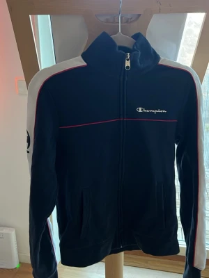 Svart ziptröja från Champion - Säljer en svart ziptröja från Champion med vita och röda detaljer. Tröjan har hög krage, hel dragkedja och vita ränder längs ärmarna samt Champion-logga på bröstet och ärmen. Perfekt för en sportig och avslappnad stil. Väldigt liten i storlek mer XS