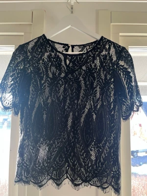 Svart spetsblus med kort ärm - Säljer en svart blus i spets med korta ärmar och rund hals. Blusen har ett detaljerat spetsmönster och fina fransiga kanter nedtill och vid ärmsluten. Perfekt att styla med jeans eller kjol för en trendig look.