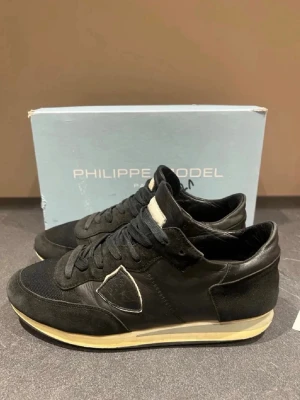 Svarta Philippe Model sneakers - Stilrena svarta sneakers från Philippe Model med klassisk låg siluett. Skorna har ovandel i mocka och mesh, detaljer i läder och vit sula med beige inslag. Ikonisk sköldlogga på sidan och svart snörning. Perfekta för dig som gillar en clean och tidlös look. Skorna är storlek 41 men passar 40 och 42. Skick 7/10. Priset kan diskuteras!