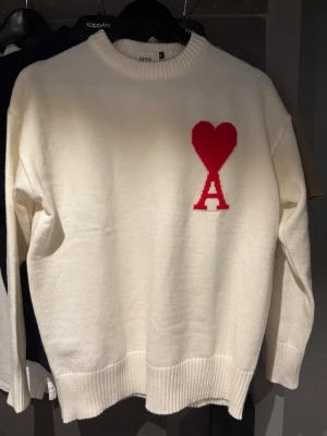 Vit stickad tröja från Ami Paris - Vit stickad tröja från Ami Paris med röd hjärtlogga och bokstaven A på bröstet. Tröjan har rund halsringning, ribbade muddar och en avslappnad passform. Perfekt för dig som gillar stilrena plagg med en twist. 
