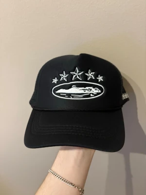 Corteiz Keps - Snygg svart truckerkeps med vit brodyr framtill som visar tre stjärnor och ett motiv av en bil. På sidan finns fler broderade stjärnor. Kepsen har mesh på sidorna och justerbar snapback. Perfekt för dig som gillar streetwear och vill sticka ut.