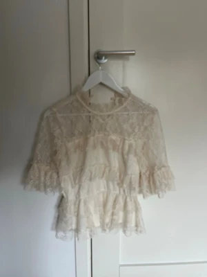 Vit spetsblus med volanger - Romantisk vit spetsblus med volanger och hög hals. Blusen har trekvartsärm och är tillverkad i ett tunt, genombrutet spetsmaterial med fina detaljer. Perfekt för dig som gillar en feminin och trendig stil.