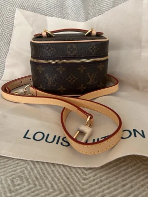  Monogram handväska - Säljer en liten och trendig handväska  sminkväska i klassiskt brunt monogrammönster. Väskan har guldfärgade detaljer, beige läderrem och dragkedja. Perfekt crossbody-modell med justerbar axelrem och snygg topphandtag. Äkta skinn och canvas. 