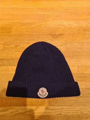 Mörkblå ribbstickad mössa Moncler - Snygg mörkblå ribbstickad mössa från Moncler med uppvikt kant och klassisk Moncler-logga framtill. Tillverkad i mjukt material som håller dig varm och ger en clean look. Perfekt accessoar för kalla dagar.