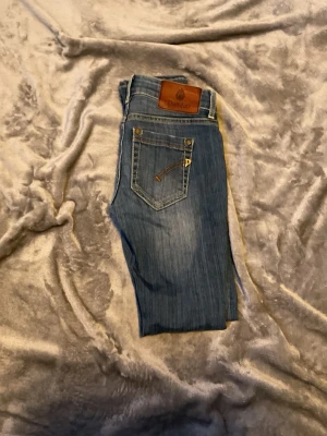 Blå smala jeans från Dovilo - Säljer ett par blåa jeans från Dondup med klassisk femficksdesign och snygga kontrastsömmar. Jeansen har en brun patch med logga bak i midjan och är tillverkade i ett mjukt denimtyg som sitter skönt. Perfekta för dig som gillar en enkel men stilrena look.