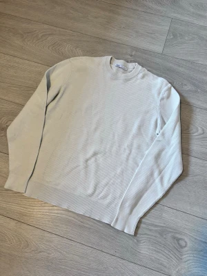 Beige stickad tröja från Zara - Snygg beige stickad tröja från Zara med rund halsringning och diskret struktur i materialet. Tröjan har långa ärmar och ribbade muddar vid ärmslut och nederkant. Perfekt basplagg till garderoben.
