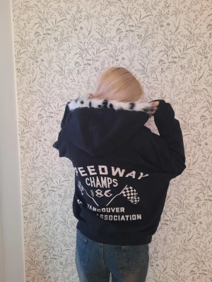 Svart hoodie med tryck och pälsdetalj - Cool svart hoodie med vitt tryck på ryggen och framsidan, samt en unik huva med svartvit fuskpäls. Påsytt för hand. Trycket har racingtema med flaggor och texten 'Speedway Champs 1986 Vancouver Association'. Perfekt för dig som gillar streetstyle.