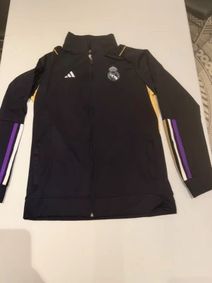 Real madrid tracksuit Adidas - Snygg svart träningskofta från Adidas med Real Madrid-logga på bröstet. Jackan har dragkedja, höga krage och detaljer i gult, vitt och lila längs ärmarna. Perfekt för träning eller att bära till vardags för dig som älskar fotboll.