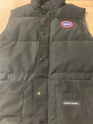 Svart dunväst från Canada Goose - Snygg svart dunväst från Canada Goose med klassisk logotyp på bröstet och två stora fickor framtill. Västen har hög krage och stängs med tryckknappar. Perfekt för kalla dagar när du vill hålla värmen och ändå se stilren ut.