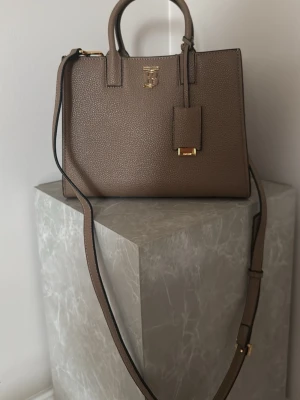 Brun Burberry handväska i läder - Snygg brun handväska från Burberry i strukturerat läder med guldfärgade detaljer och klassisk TB-logga framtill. Väskan har både handtag och justerbar axelrem, samt en dekorativ tag. Perfekt för dig som gillar stilrena accessoarer. 25 cm bred och 19cm lång 